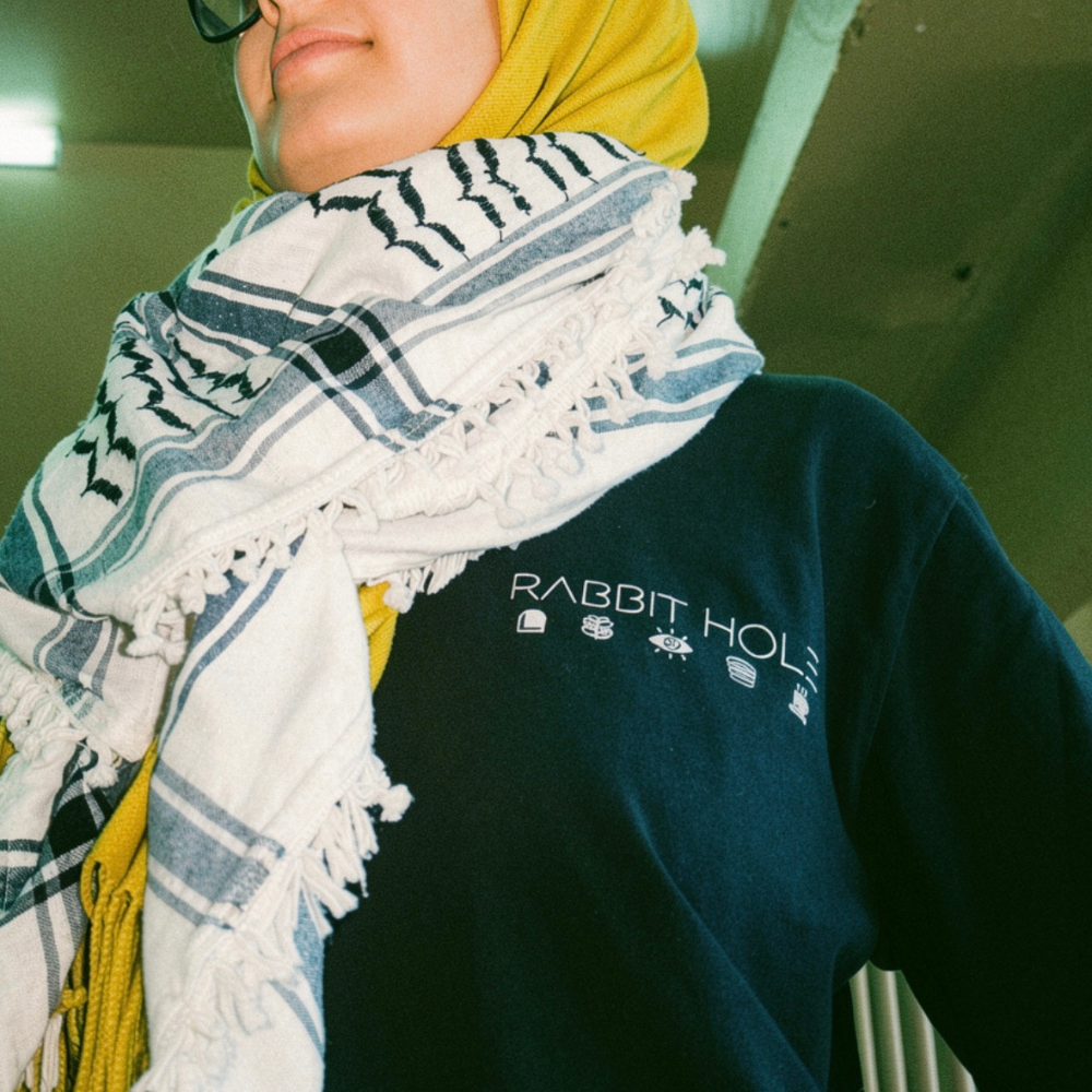 Rayyana Alkebsi is wearing a black long sleeve t-shirt with 'Rabbit Hole' branding, a white and black traditional patterned keffiyeh, and a yellow headscarf. The shirt is an on-going partnership with Decolonial clothing who produces all of Rabbit Hole's apparel. 